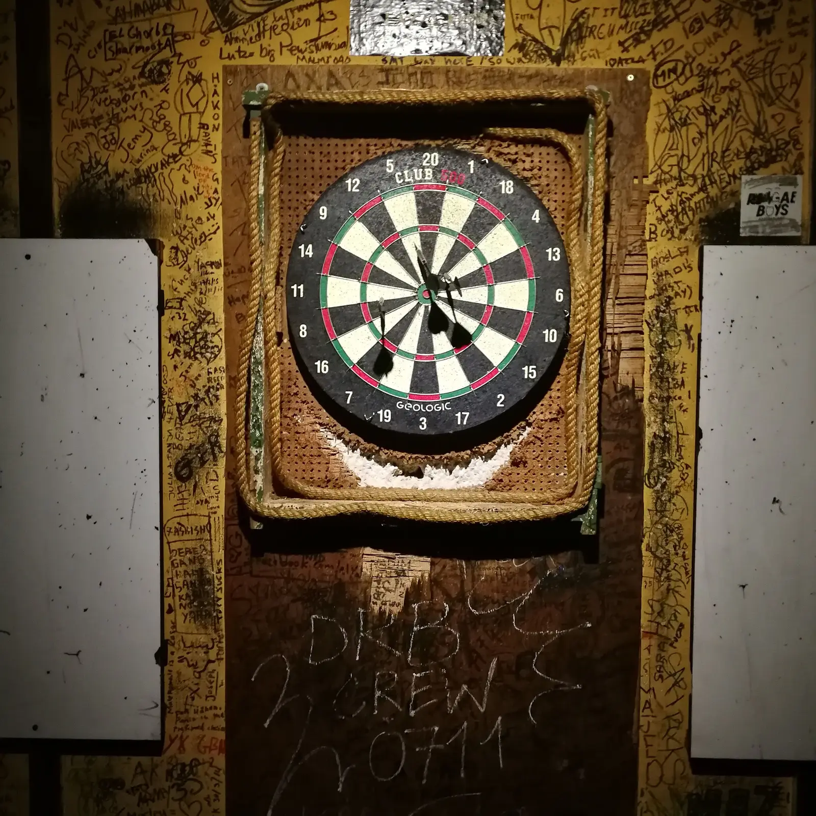 A heavily used dartboard mounted on a wooden wall covered in scrawled signatures